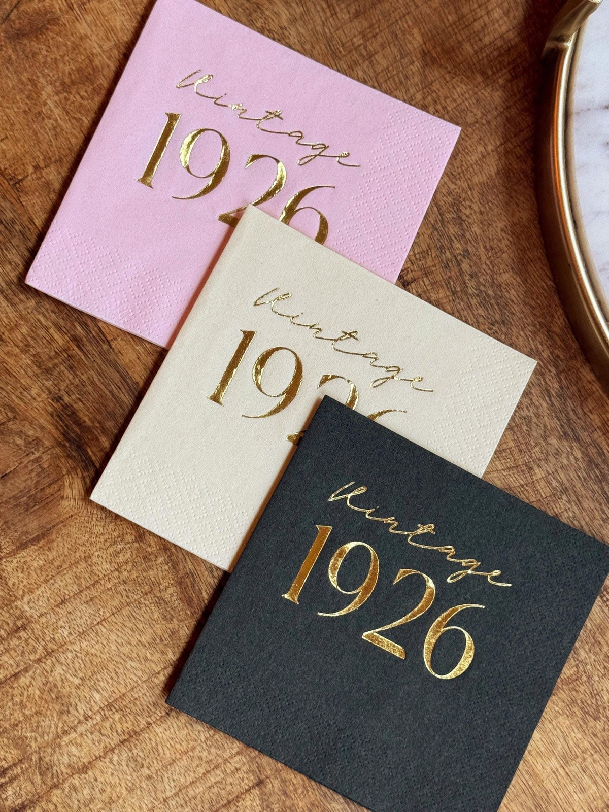 Pink, cream, and black paper napkins printed with gold foil Vintage 1926 text, laid on a wooden tabletop.