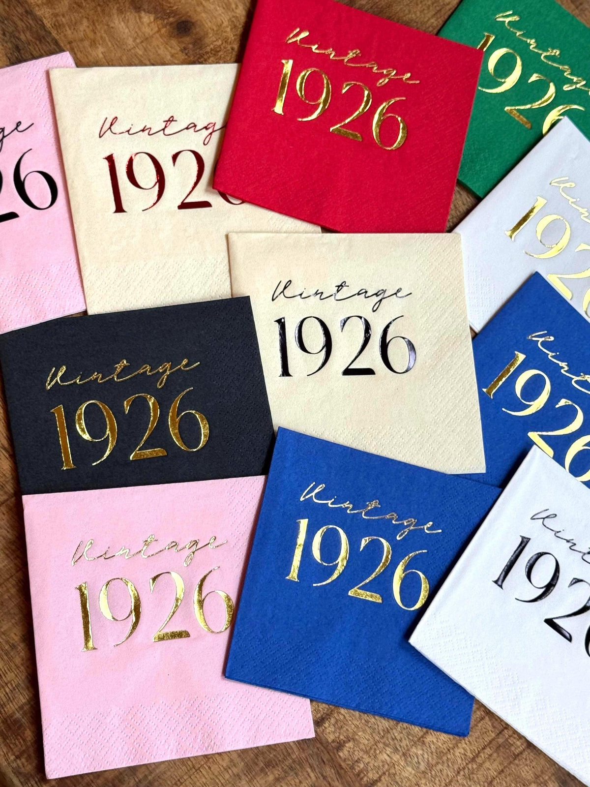 A scattering of colorful cocktail napkins featuring Vintage 1926 foil stamping in various metallic and matte colors on a wooden background.