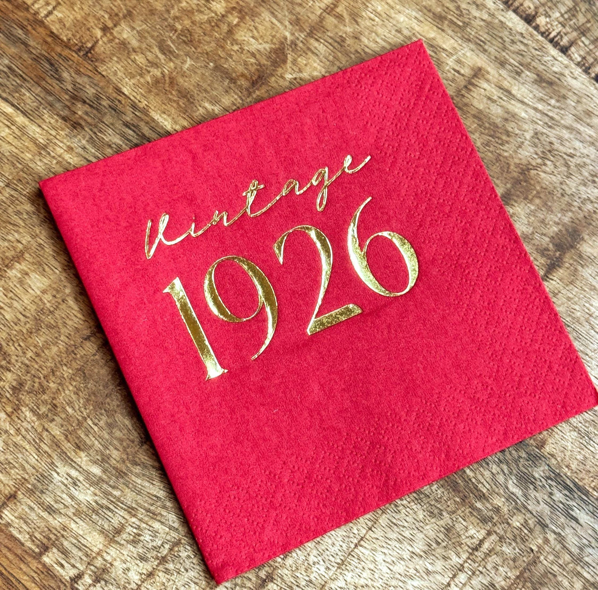 A single red cocktail napkin stamped with shiny gold Vintage 1926 lettering rests on a wooden table.