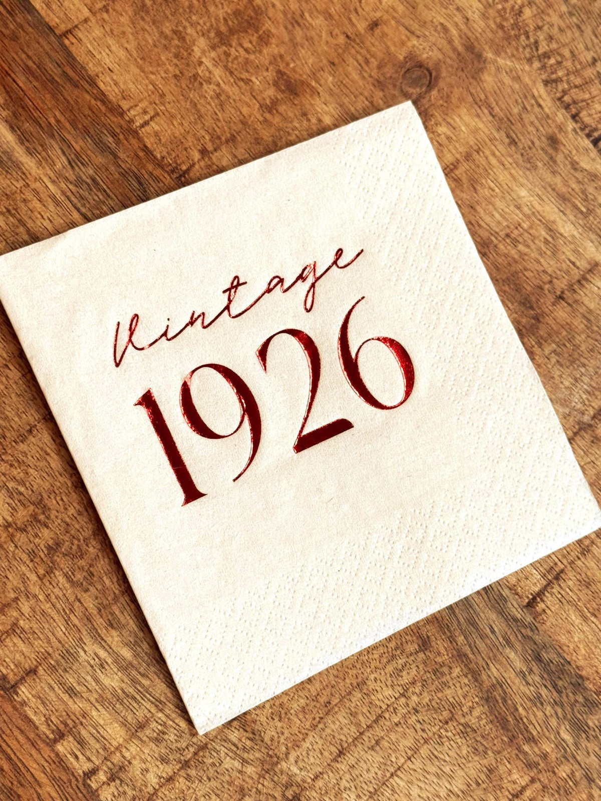 A cream-colored cocktail napkin stamped with metallic red Vintage 1926 text sits on a wooden tabletop.