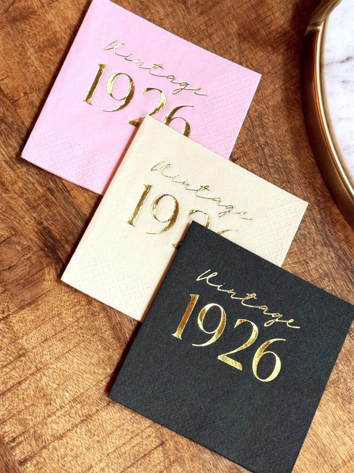 Three folded cocktail napkins in pastel pink, cream, and black are displayed on a wooden surface, each stamped with Vintage 1926 text.