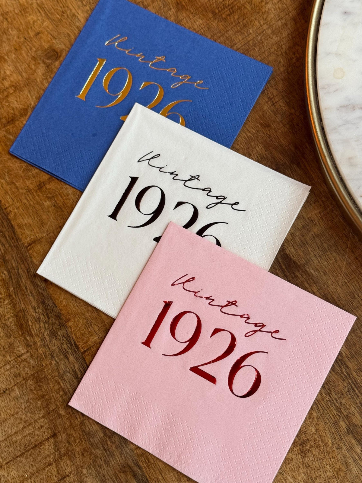 Three folded cocktail napkins in blue, white, and pink are fanned out on a wooden table, each stamped with Vintage 1926 text.