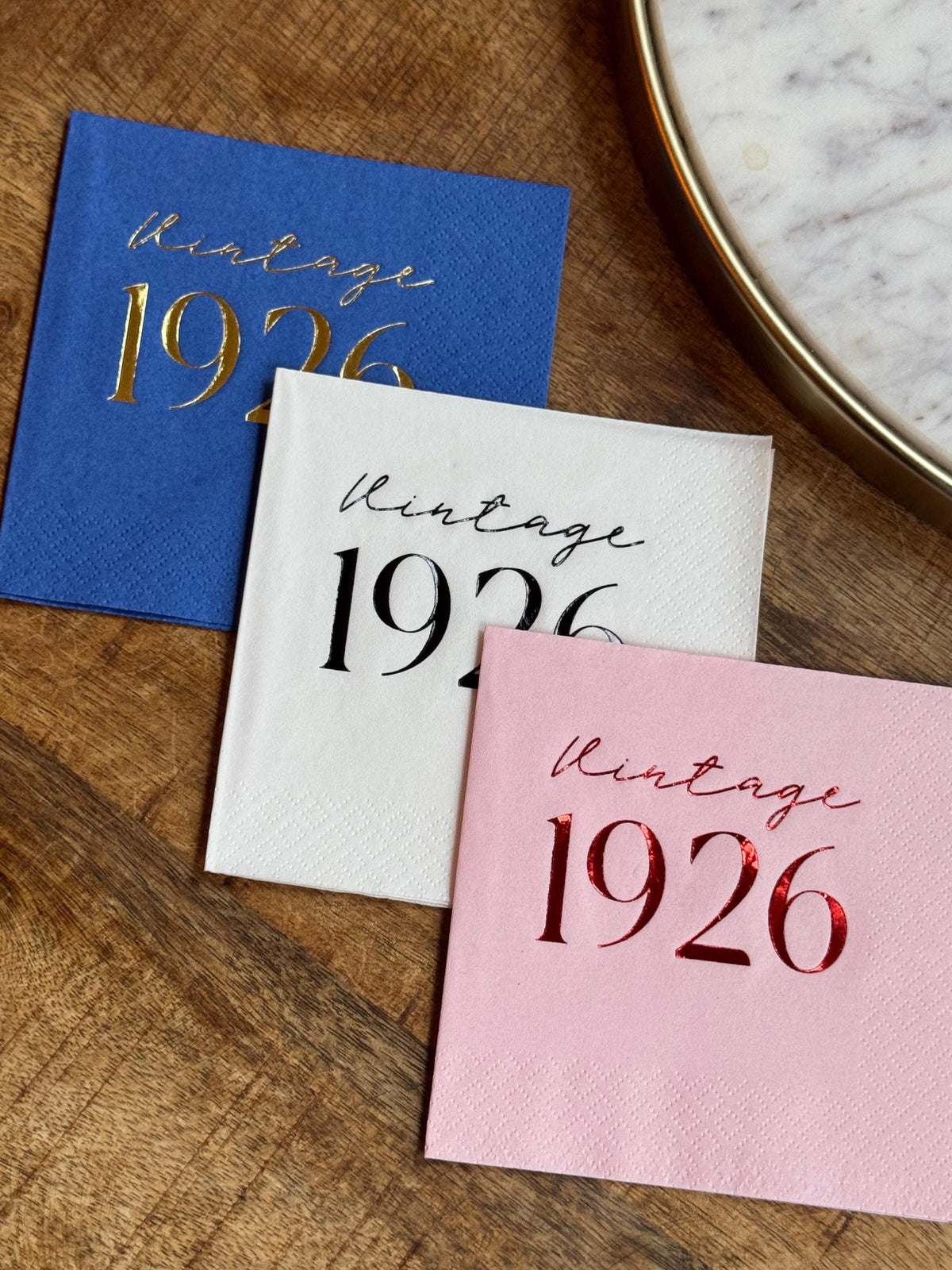 Three folded cocktail napkins in royal blue, white, and pink are arranged on a wooden surface, each featuring Vintage 1926 custom foil stamping.