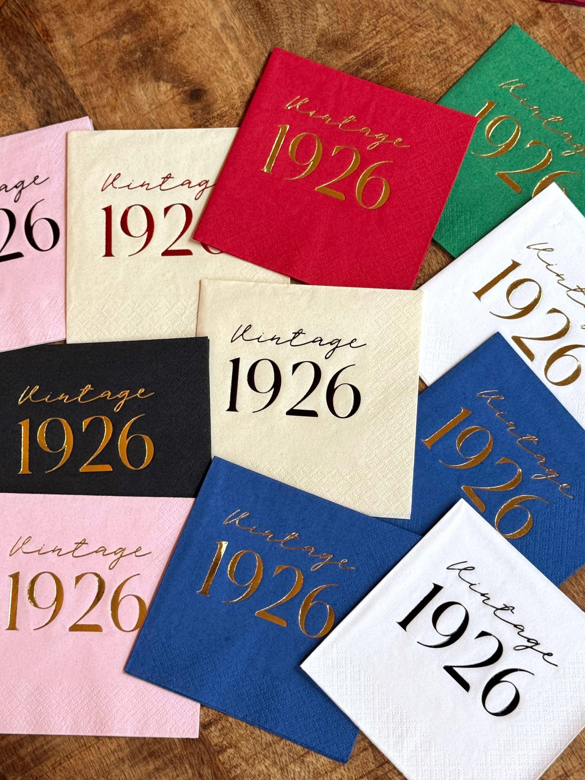 An assortment of colorful cocktail napkins stamped with Vintage 1926 text is scattered across a wooden surface.