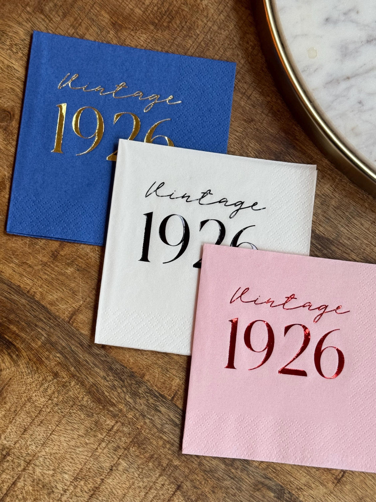 Three folded paper napkins in blue, white, and pink, printed with Vintage 1926, resting on a wooden surface beside a round tray.