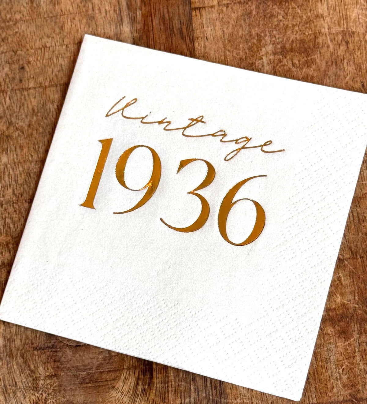 A white cocktail napkin with the words "Vintage 1936" printed in gold foil, on a rustic wooden surface.