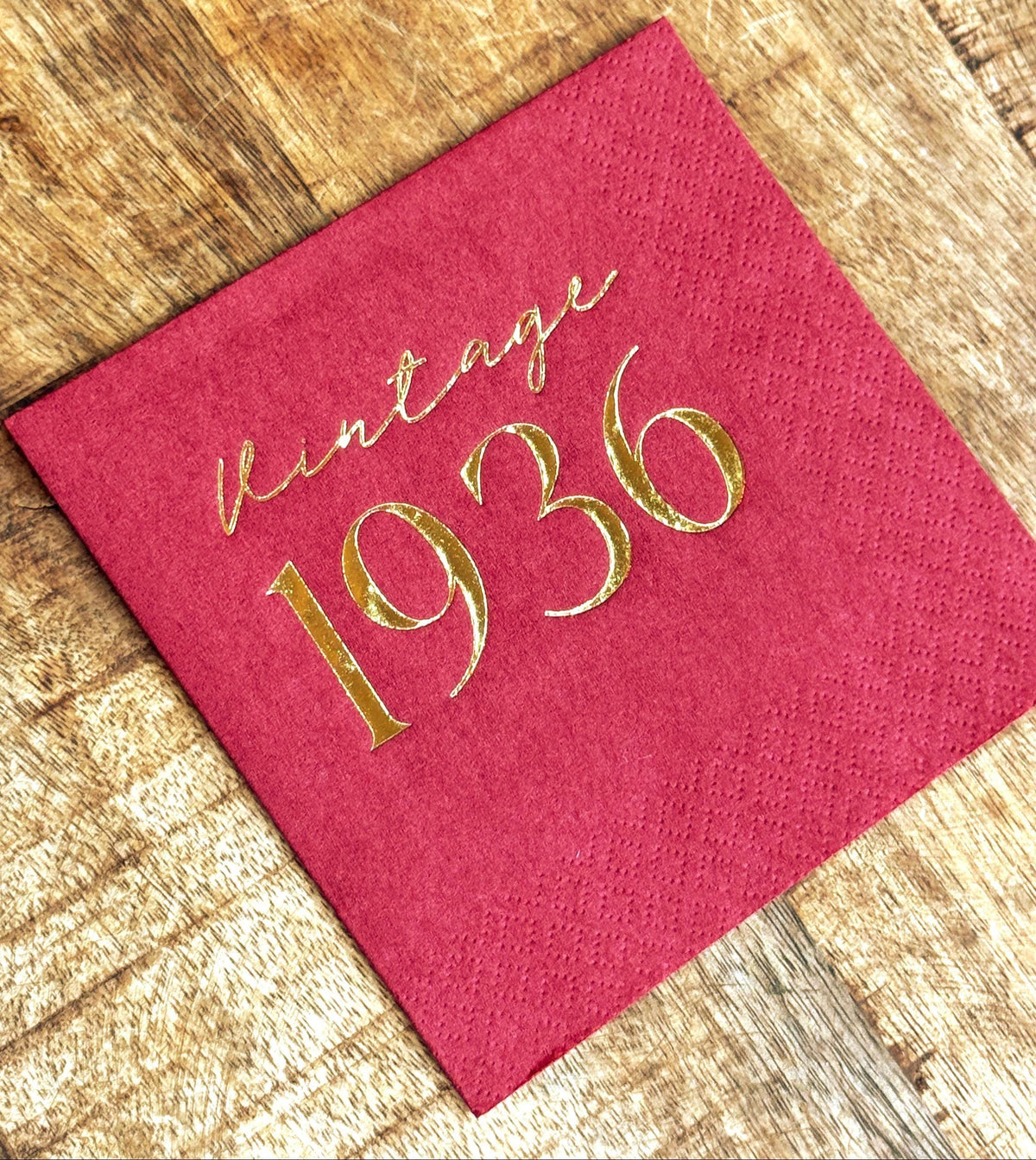 A red cocktail napkin with the words "Vintage 1936" printed in gold foil, on a rustic wooden surface.