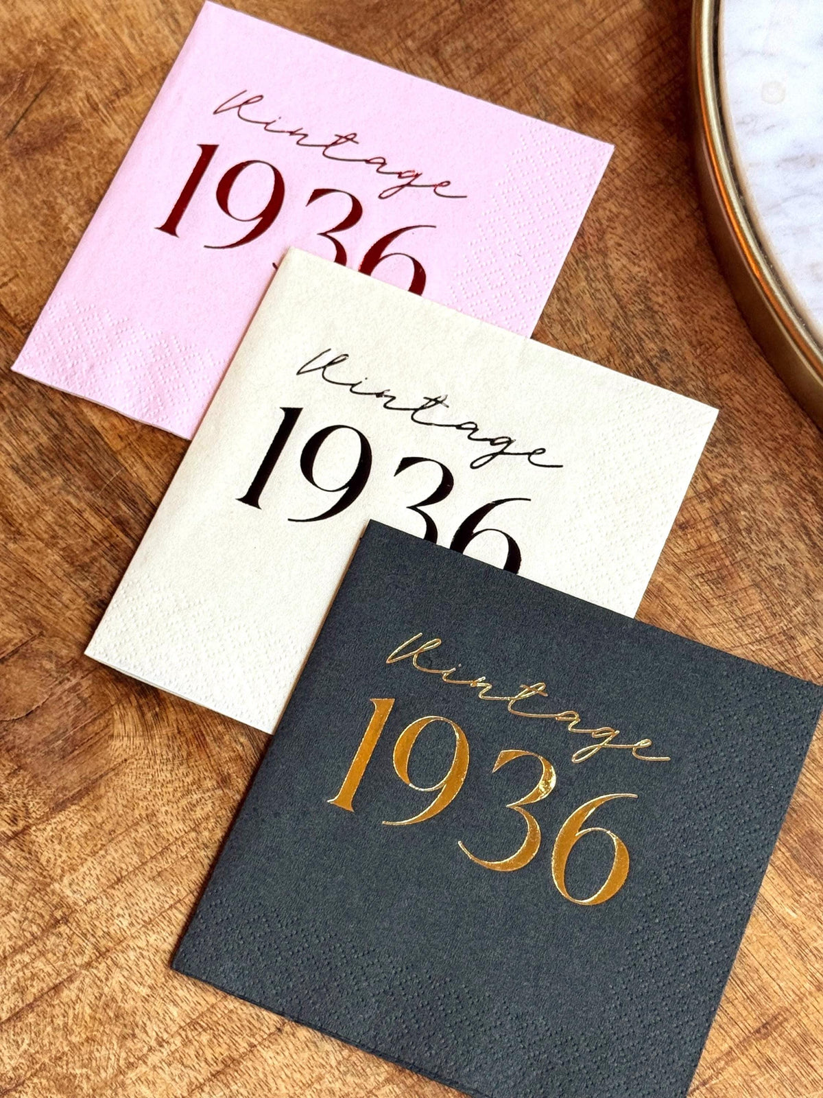 Three cocktail napkins are arranged on a wooden surface: a light pink napkin with black text, a cream napkin with black text, and a dark gray napkin with gold text, all reading "Vintage 1936".