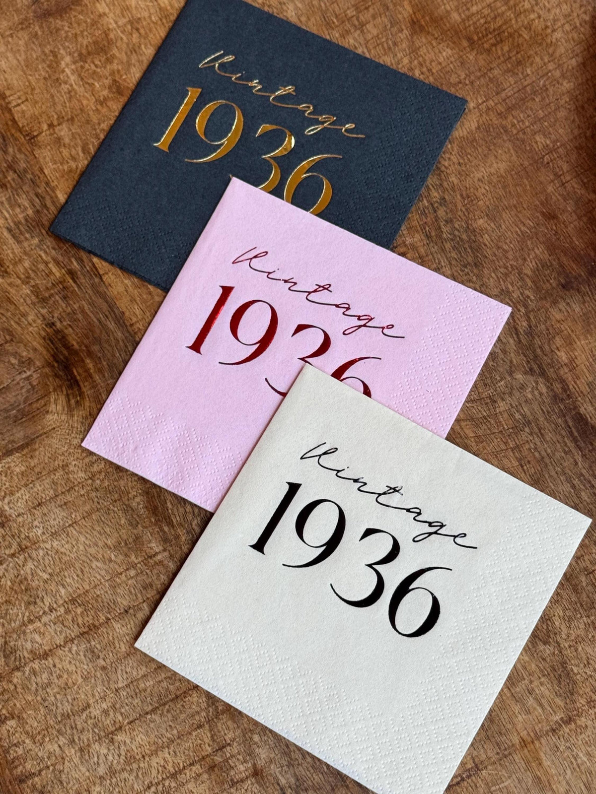 Three cocktail napkins are arranged on a wooden surface: a dark gray napkin with gold text, a light pink napkin with red text, and a cream napkin with black text, all reading "Vintage 1936".