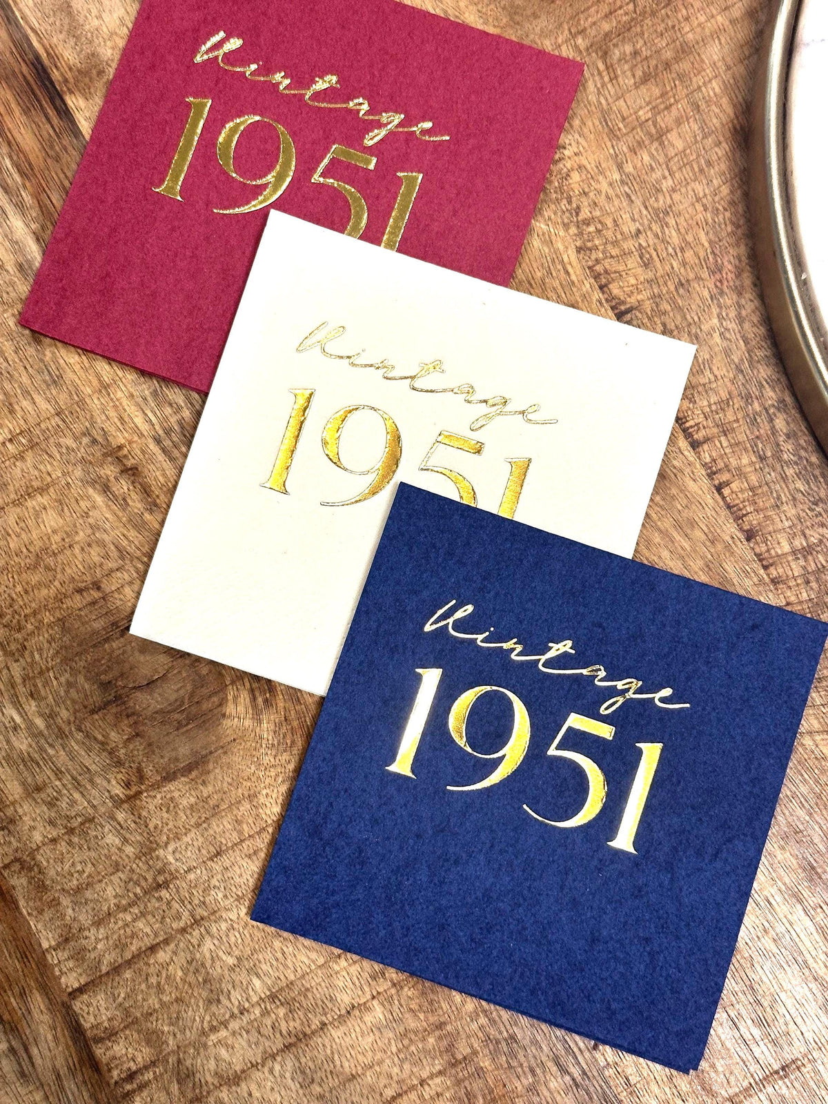 Three square paper napkins in maroon, white, and dark blue are arranged on a wooden surface, each featuring gold foil text that reads vintage 1951.