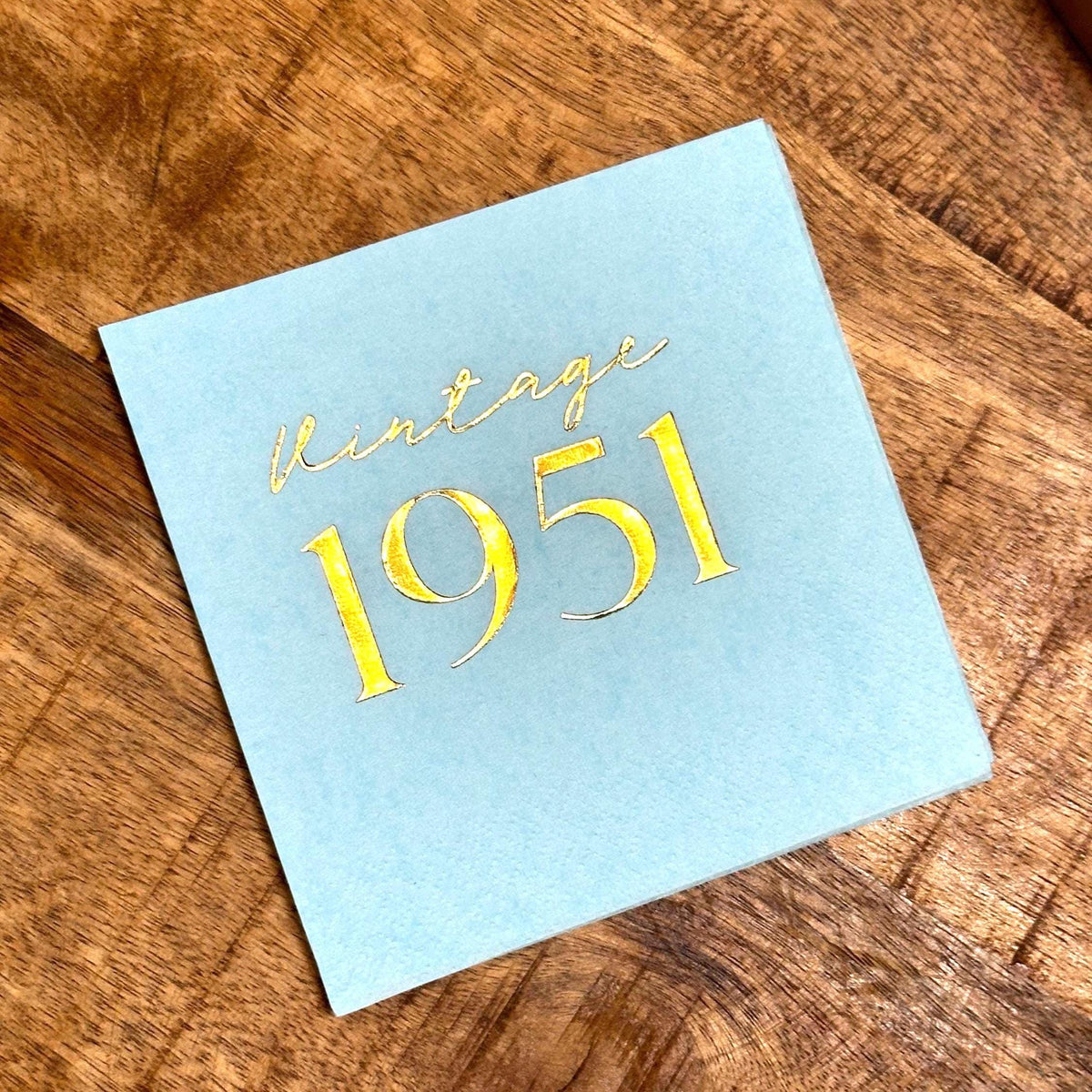 A single light blue square paper napkin rests on a wooden surface, stamped with gold foil text that reads vintage 1951.