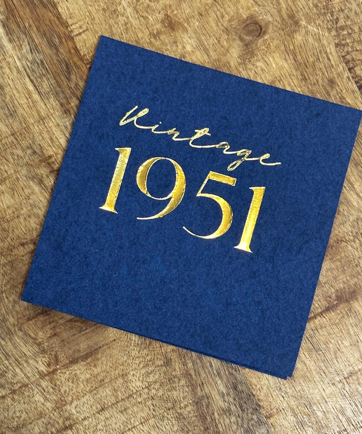 A single dark blue square paper napkin sits on a wooden surface, printed with gold foil text reading vintage 1951.
