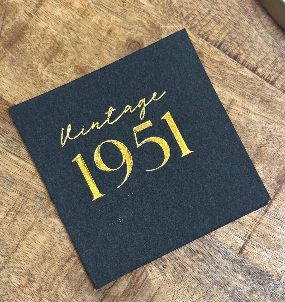A single black square paper napkin lies on a wooden surface, featuring gold foil lettering that reads vintage 1951.
