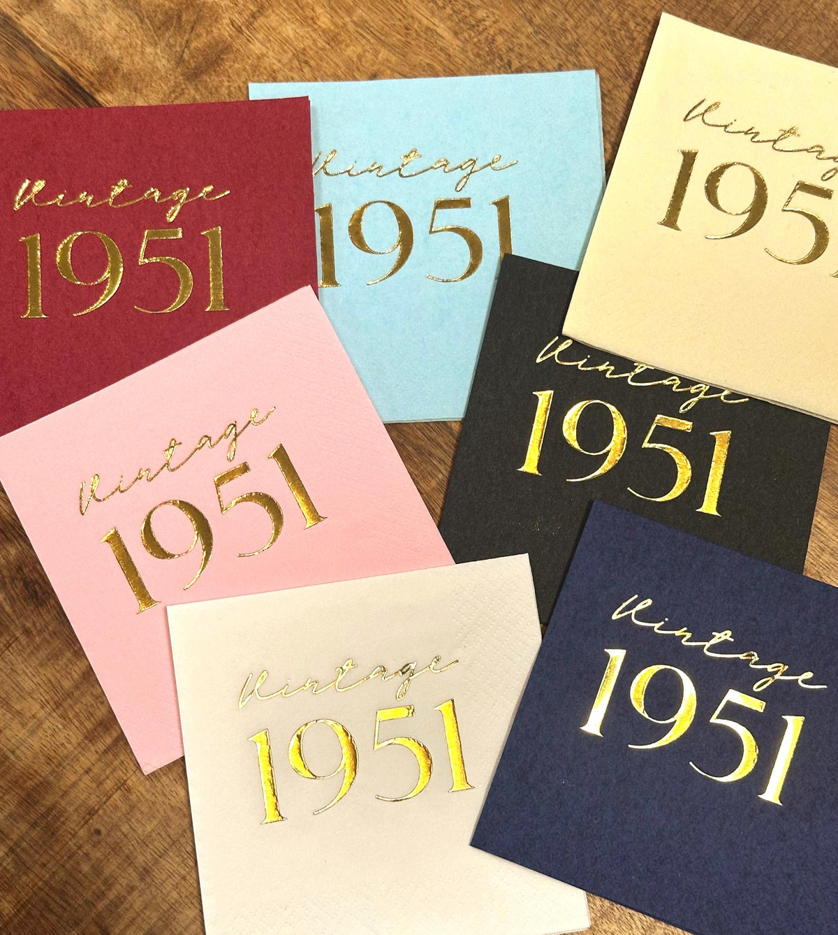 Seven square paper napkins in various colors including maroon, light blue, cream, pink, black, cream, and dark blue are arranged on a wooden table, all featuring gold foil vintage 1951 text.