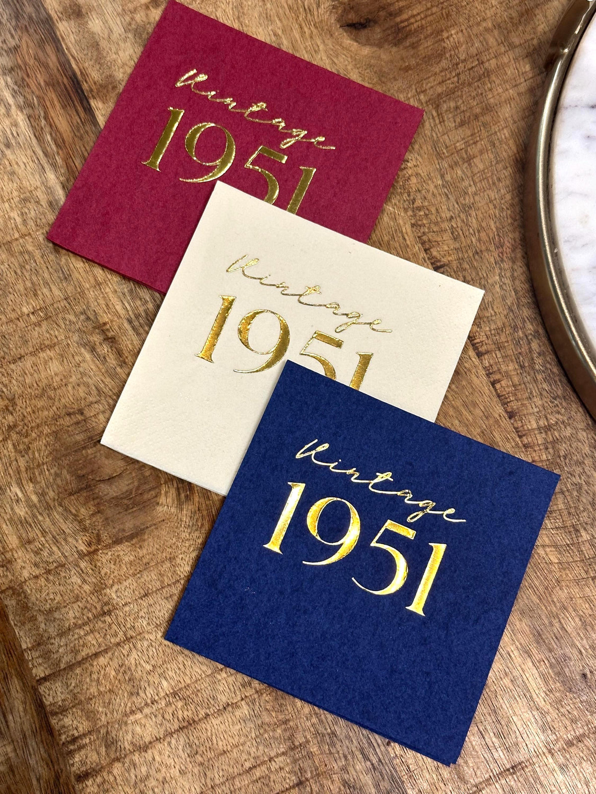Three square paper napkins in maroon, cream, and dark blue are stacked diagonally on a wooden table, each stamped with gold foil vintage 1951 text.