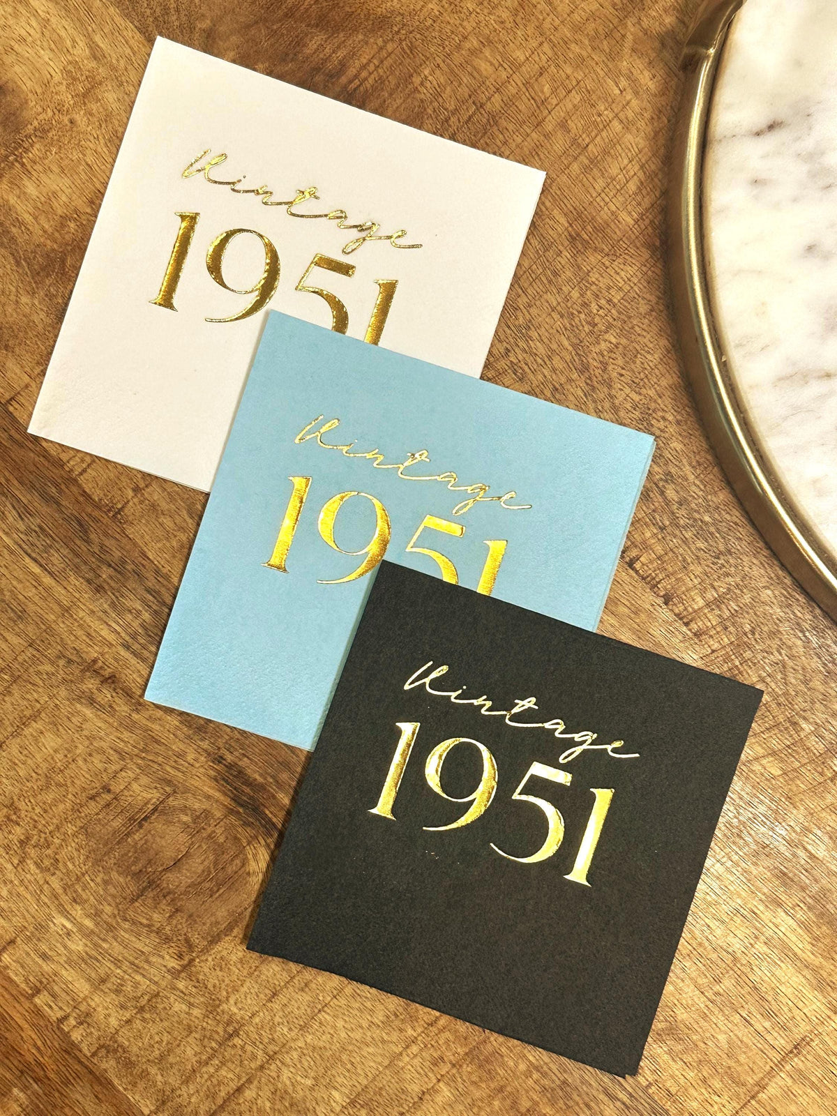 Three square paper napkins in white, light blue, and black are displayed on a wooden surface, each featuring gold foil lettering that reads vintage 1951.