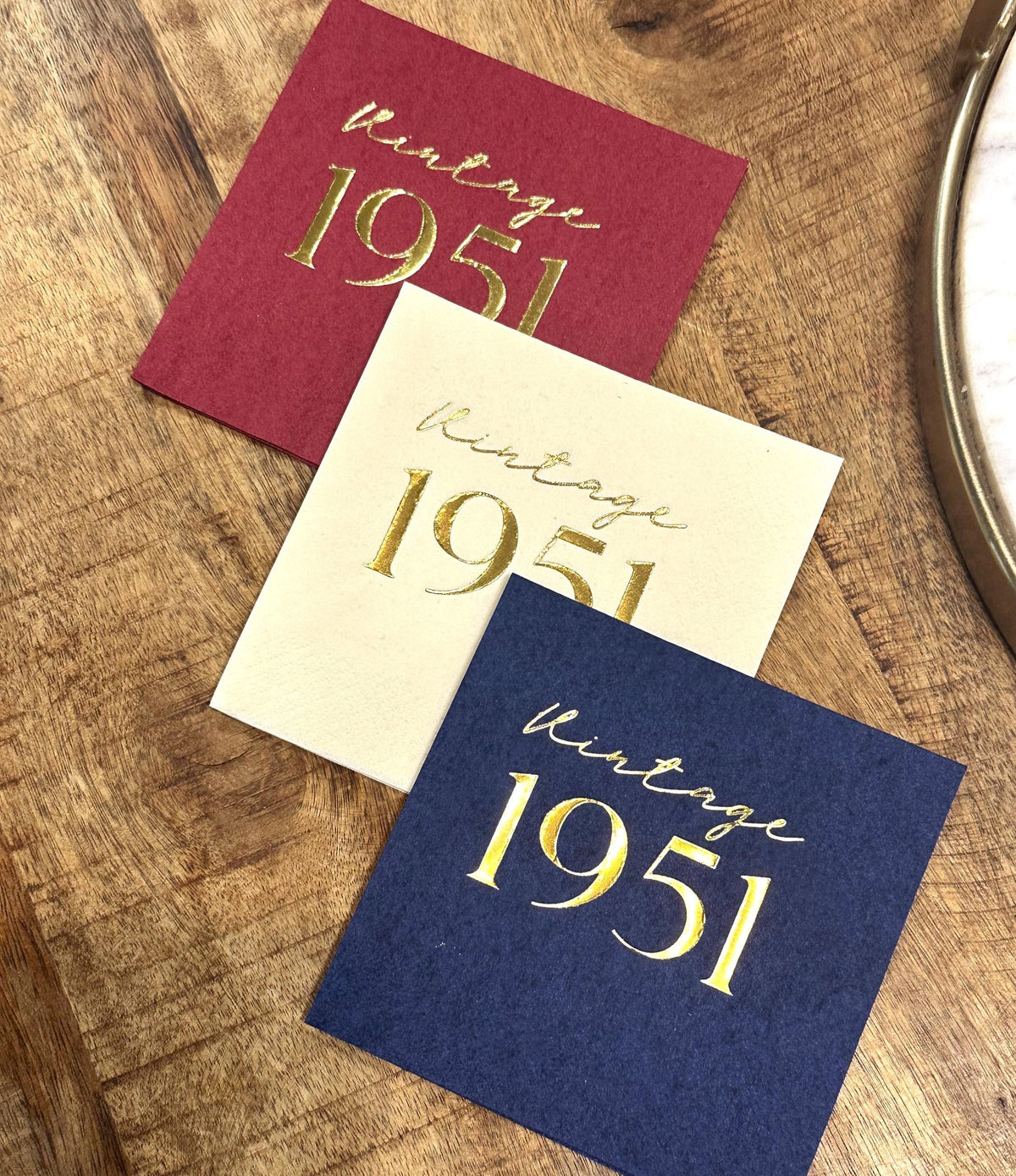 Three square paper napkins in maroon, cream, and dark blue are laid out on a wooden surface, printed with gold foil vintage 1951 text.