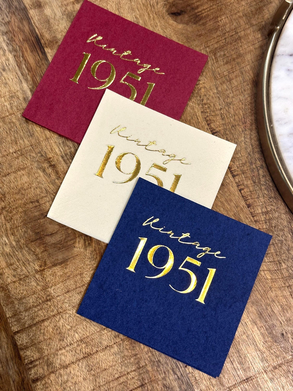 Three square paper napkins in maroon, cream, and dark blue are arranged on a wooden table, each stamped with gold foil vintage 1951 text.