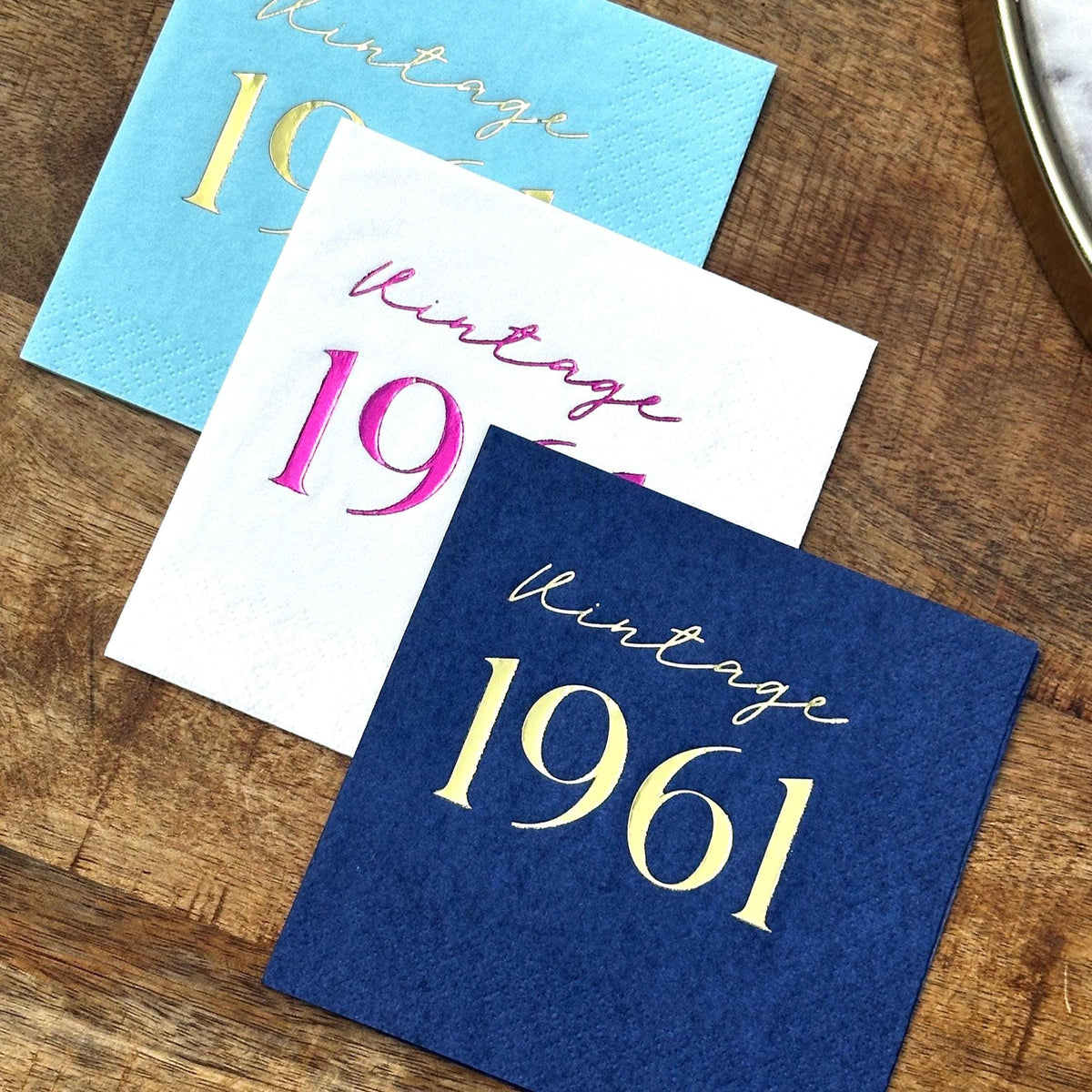 Three square cocktail napkins stacked diagonally on a wooden surface, featuring light blue, white, and dark blue with foil text.