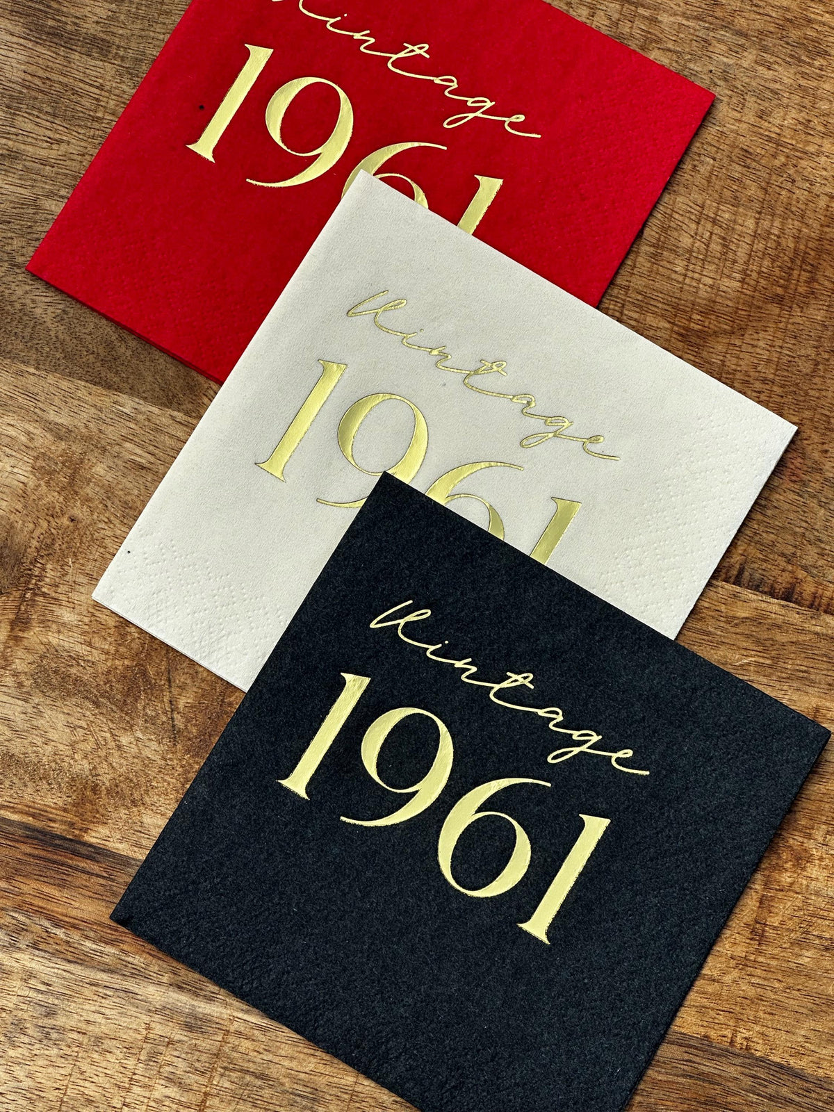 Three folded square cocktail napkins in red, cream, and black stacked diagonally on a wood surface, each foil-stamped with the text Vintage 1961 in gold.
