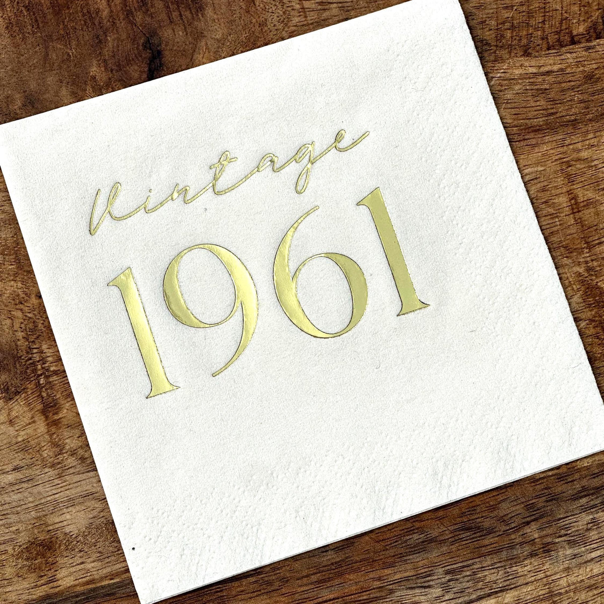 A single white square cocktail napkin with gold foil stamping that reads Vintage 1961 on a wooden tabletop.