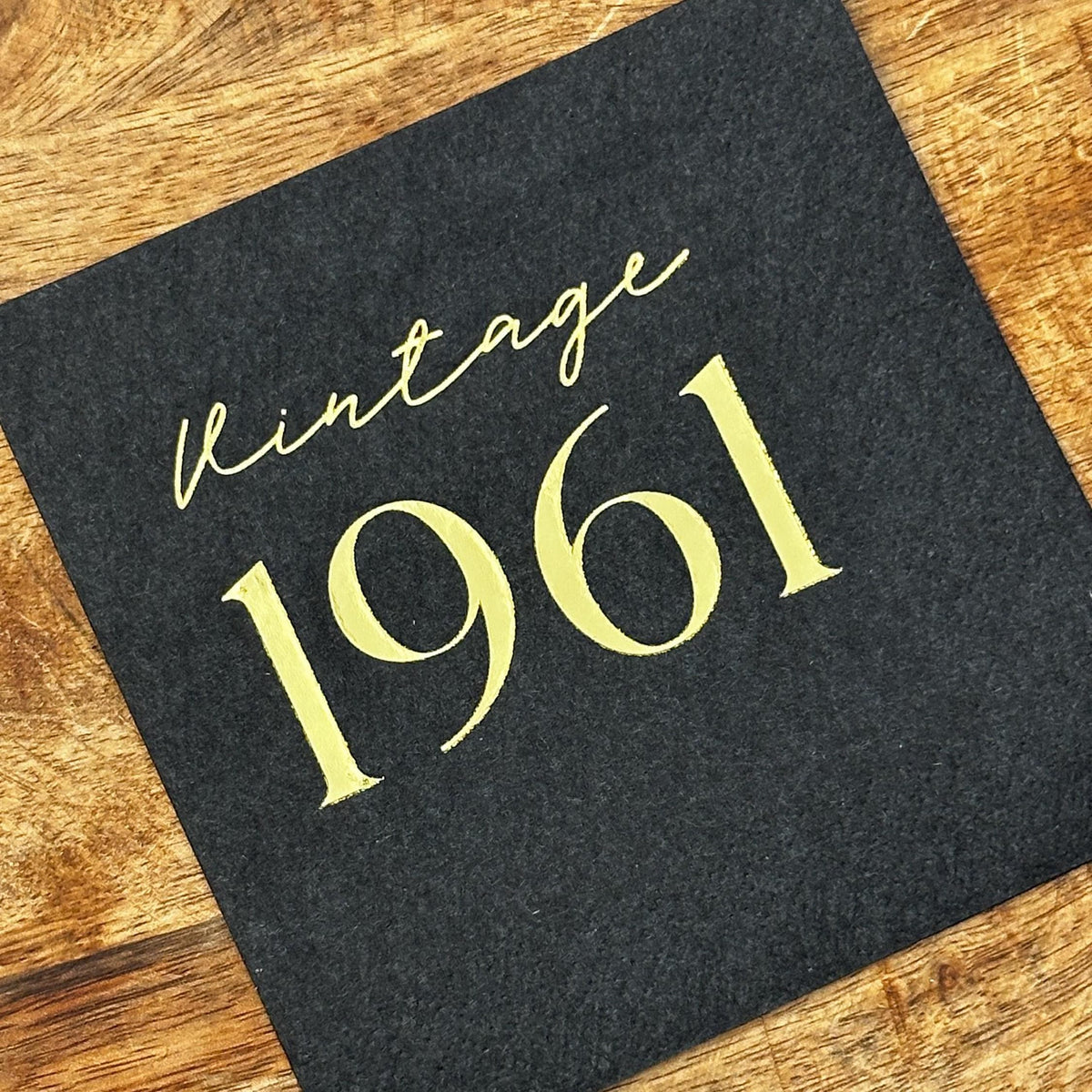 A single black square cocktail napkin printed with gold foil lettering reading Vintage 1961, set on a wood background.