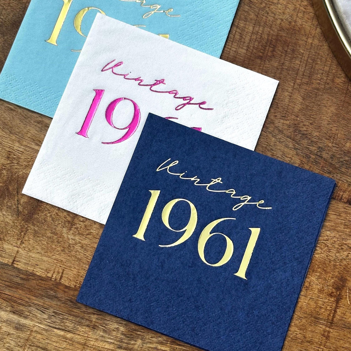 Three square cocktail napkins in light blue, white, and dark blue with metallic foil text arranged diagonally on wood.