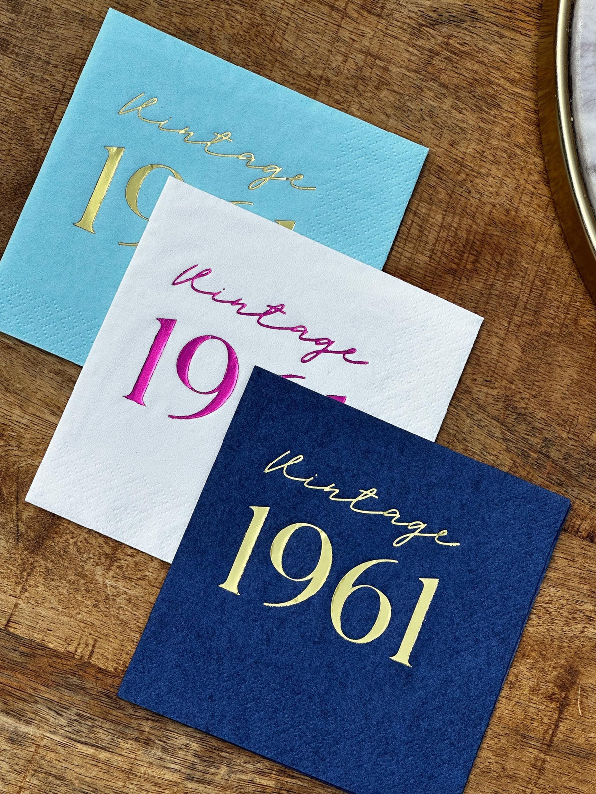Three stacked square cocktail napkins in light blue, white, and navy blue with metallic foil text on a wooden surface.