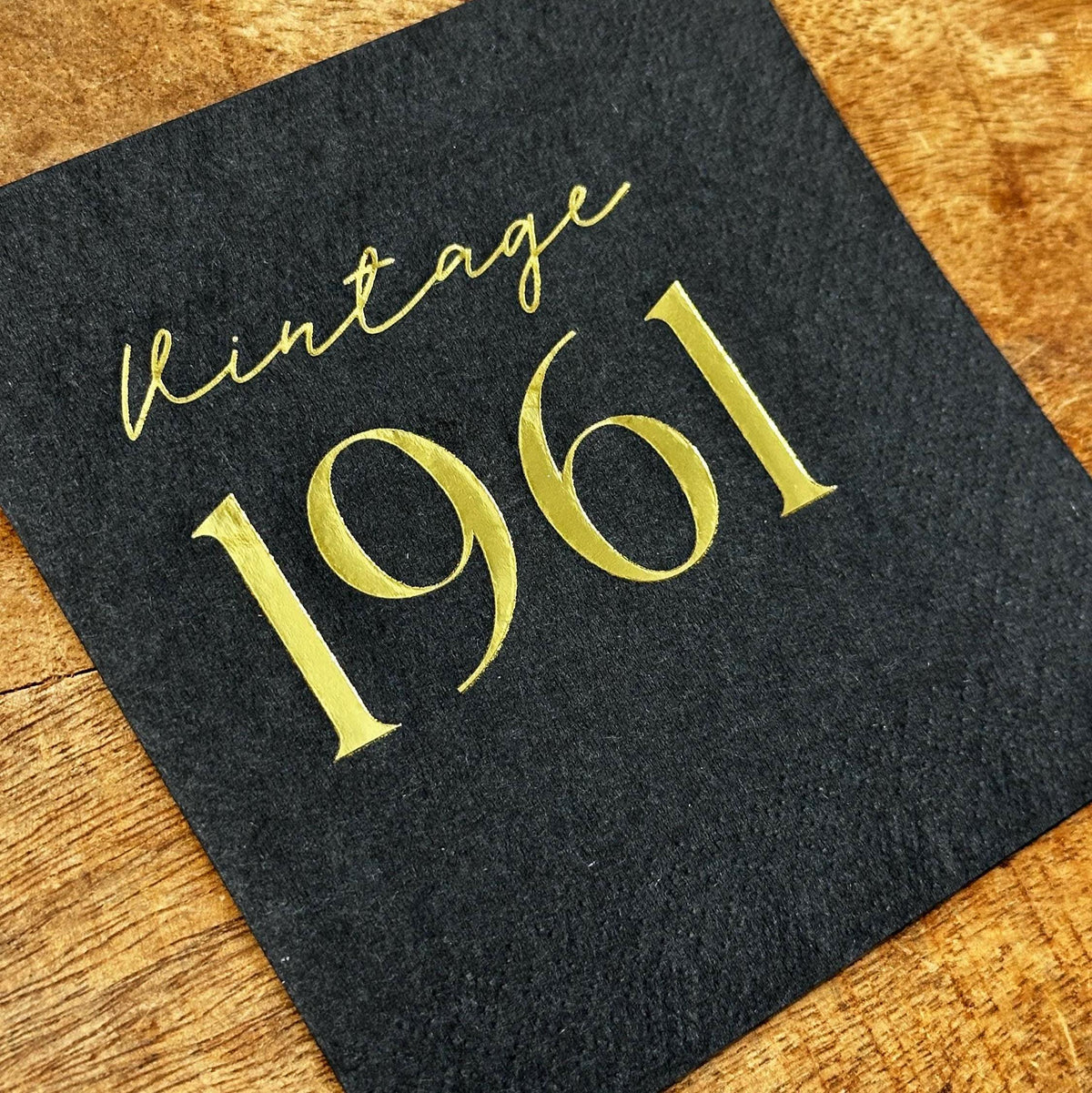 A single black square cocktail napkin with gold foil text reading Vintage 1961 placed on a wooden board.