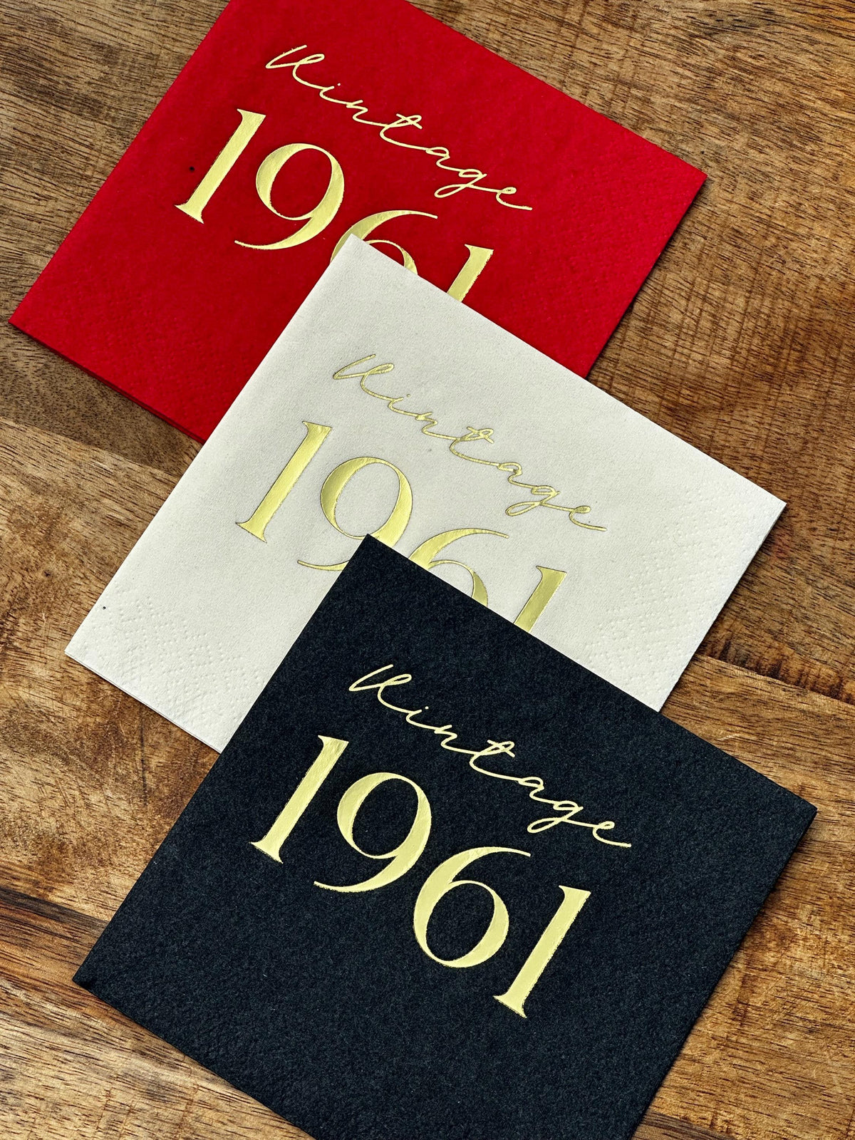 Three square cocktail napkins in red, white, and black with shiny gold foil lettering on a rustic wooden table.