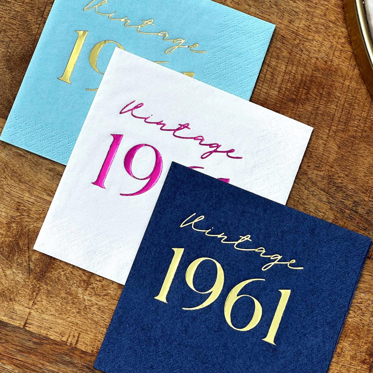 Three square cocktail napkins in light blue, white, and dark blue featuring foil text stacked on a wooden background.
