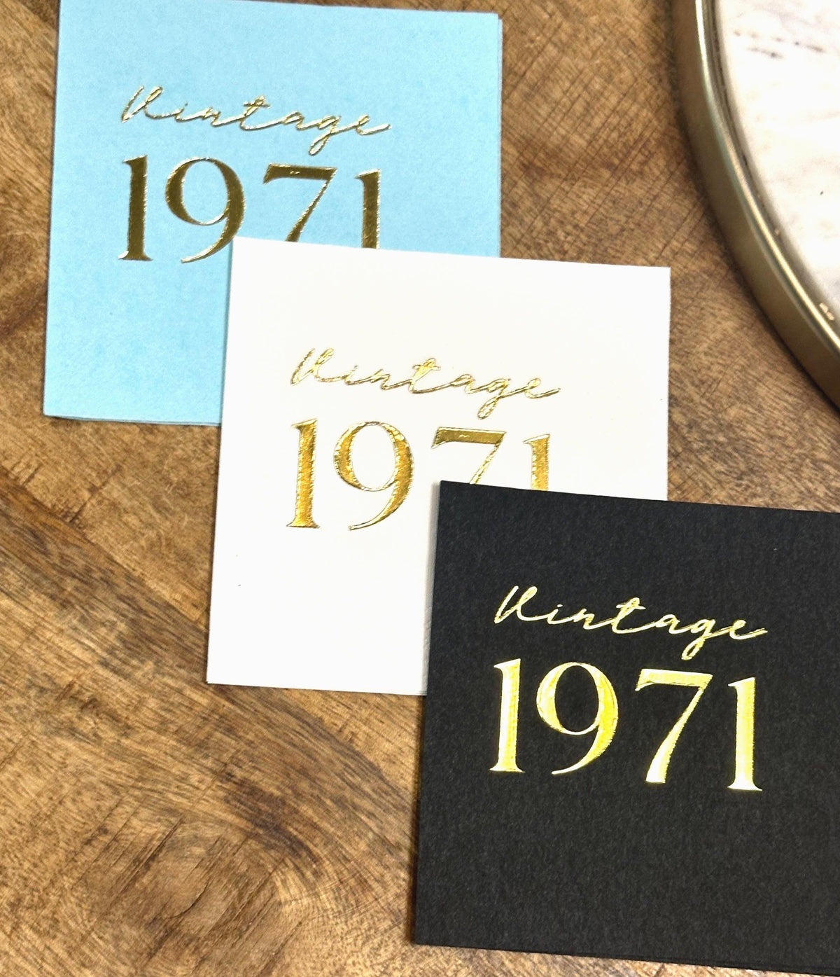 Three square napkins, light blue, white, and black, each foil-stamped with "Vintage 1971" in gold, on a wooden surface.