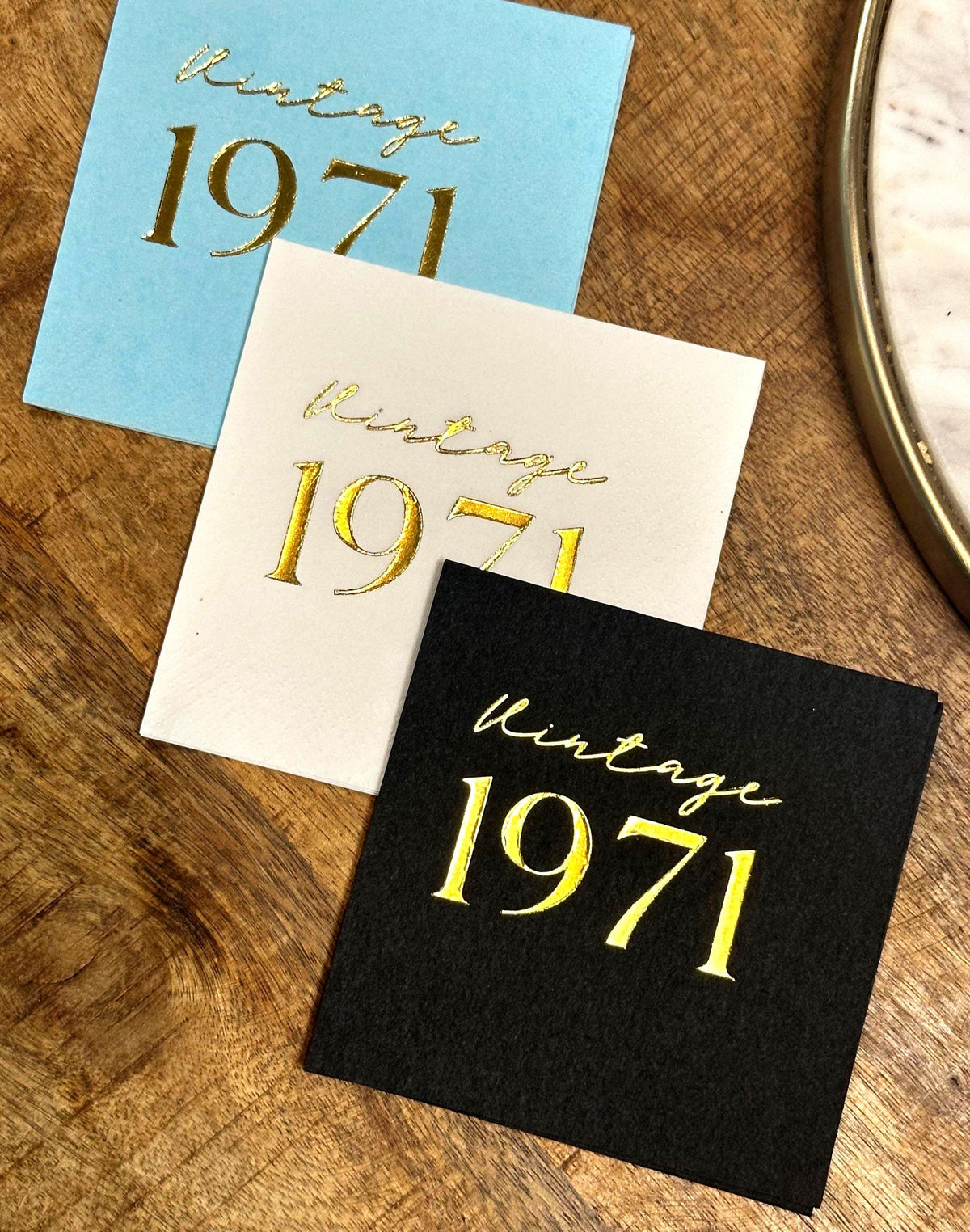 Three square napkins, light blue, off-white, and black, each foil-stamped with "Vintage 1971" in gold, on a wooden surface.