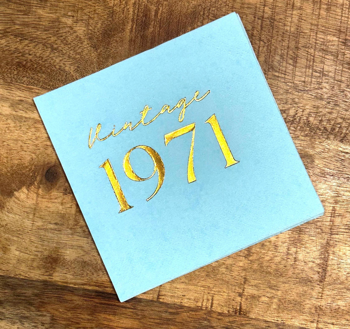 A single light blue square napkin, foil-stamped with "Vintage 1971" in gold, on a wooden surface.