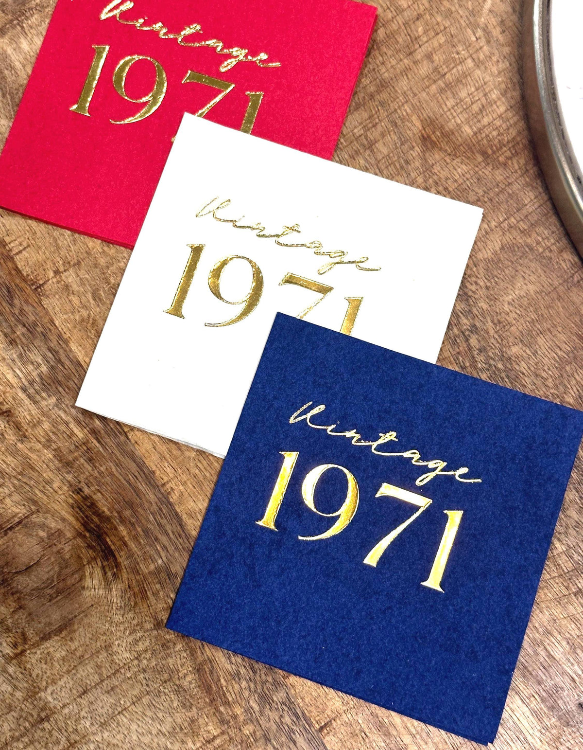Three square napkins, red, white, and navy blue, each foil-stamped with "Vintage 1971" in gold, on a wooden surface.
