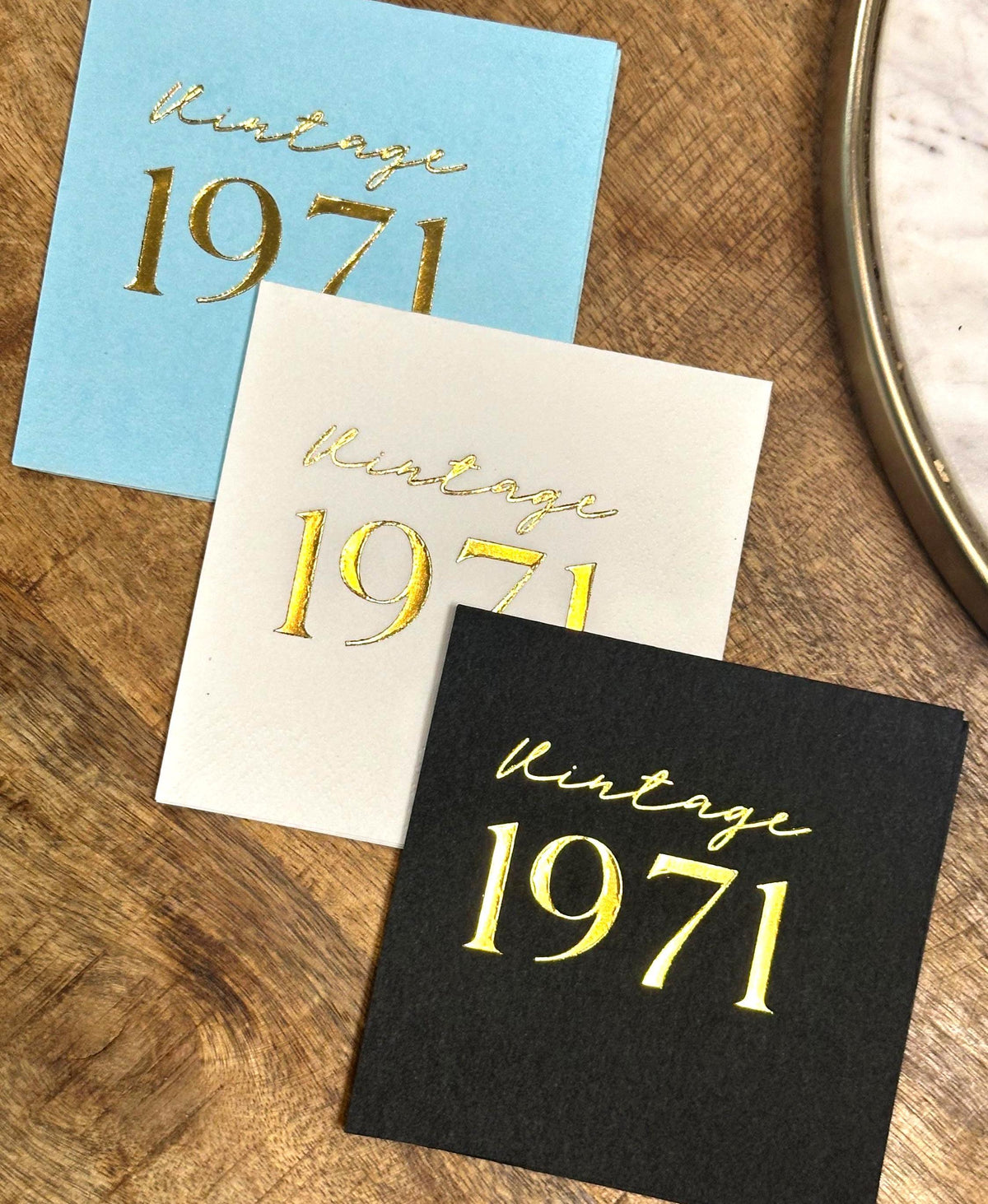 Three square napkins, light blue, off-white, and black, each foil-stamped with "Vintage 1971" in gold, on a wooden surface.