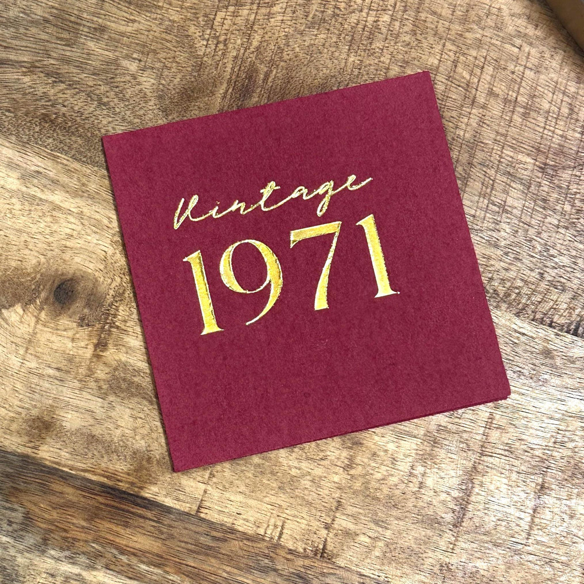 A single maroon square napkin, foil-stamped with "Vintage 1971" in gold, on a wooden surface.