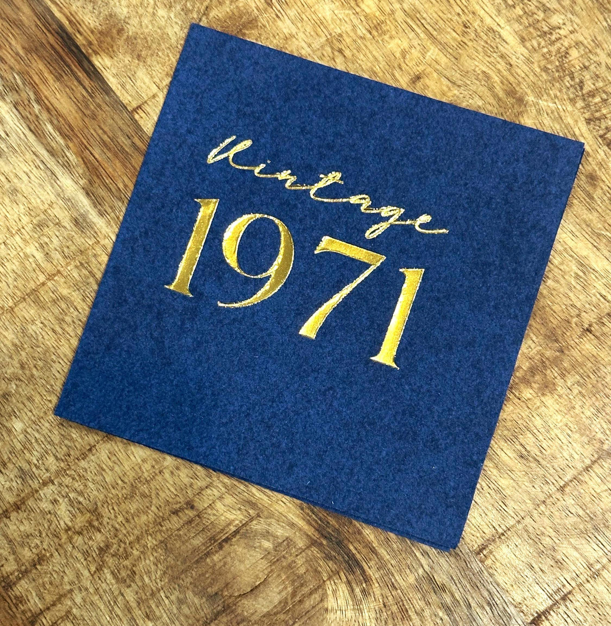 A single navy blue square napkin, foil-stamped with "Vintage 1971" in gold, on a wooden surface.