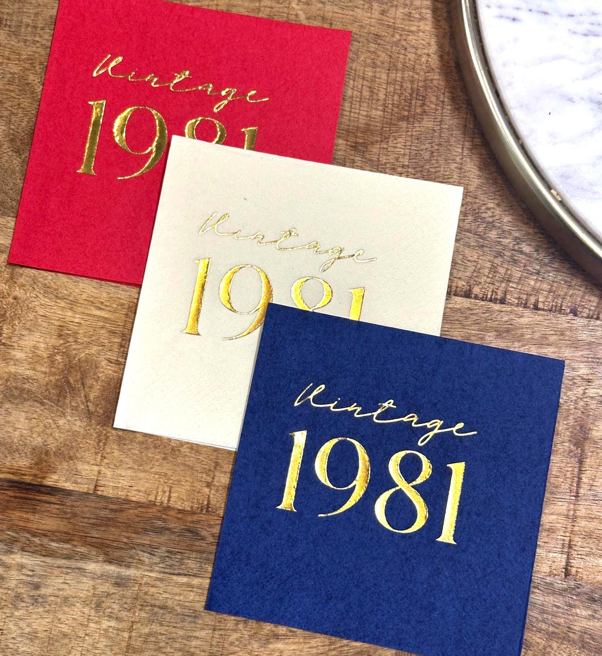 Three square paper napkins in red, cream, and dark blue, each stamped with the text Vintage 1981 in gold foil.