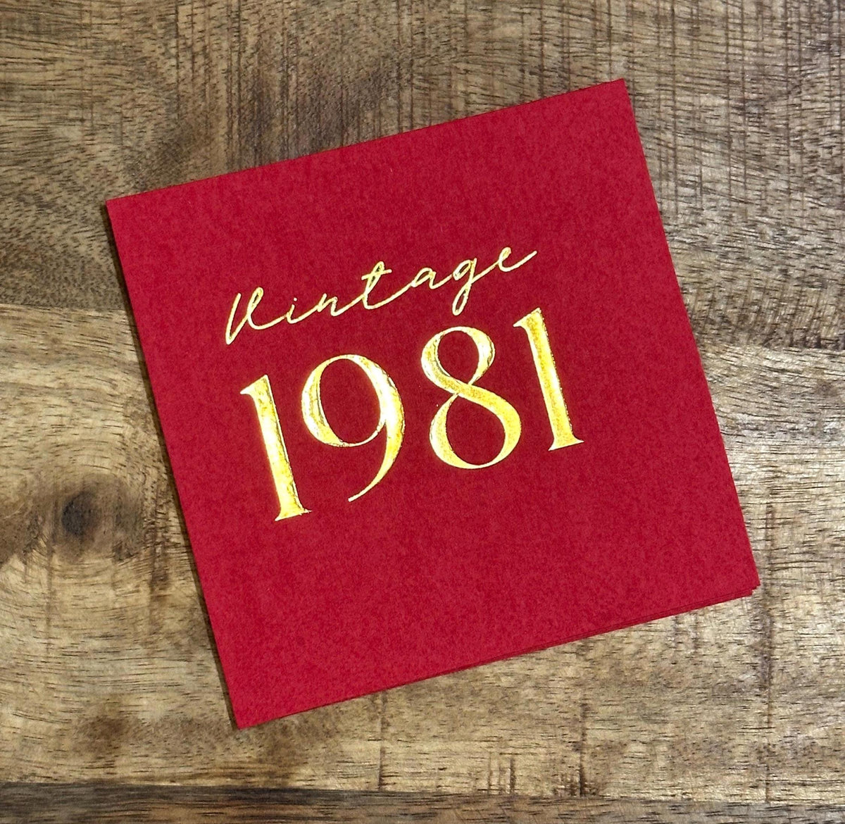 A single square red paper napkin stamped with the text Vintage 1981 in gold foil, resting on a wooden surface.