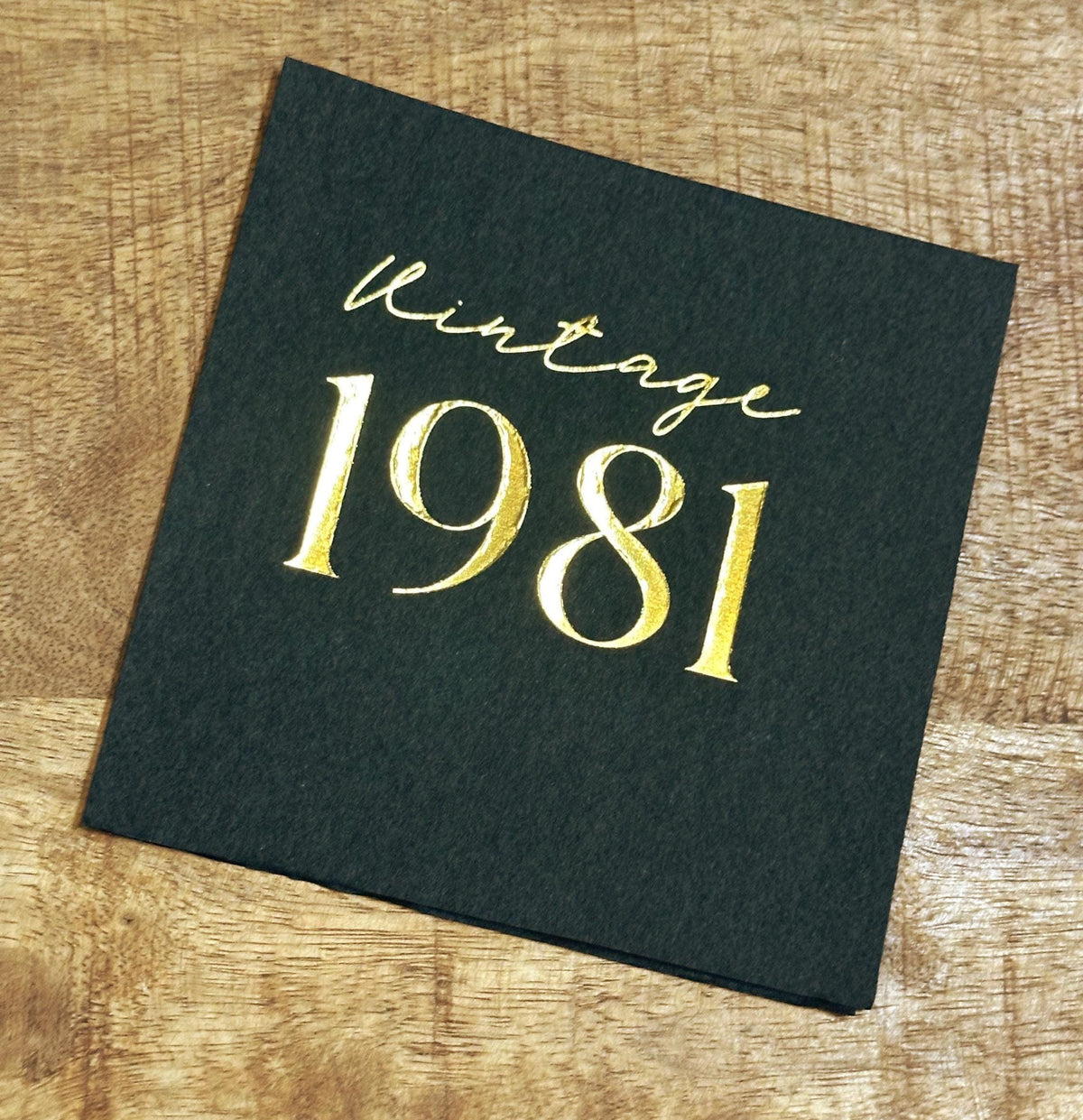 A single square dark gray paper napkin stamped with the text Vintage 1981 in gold foil, placed on a wooden tabletop.