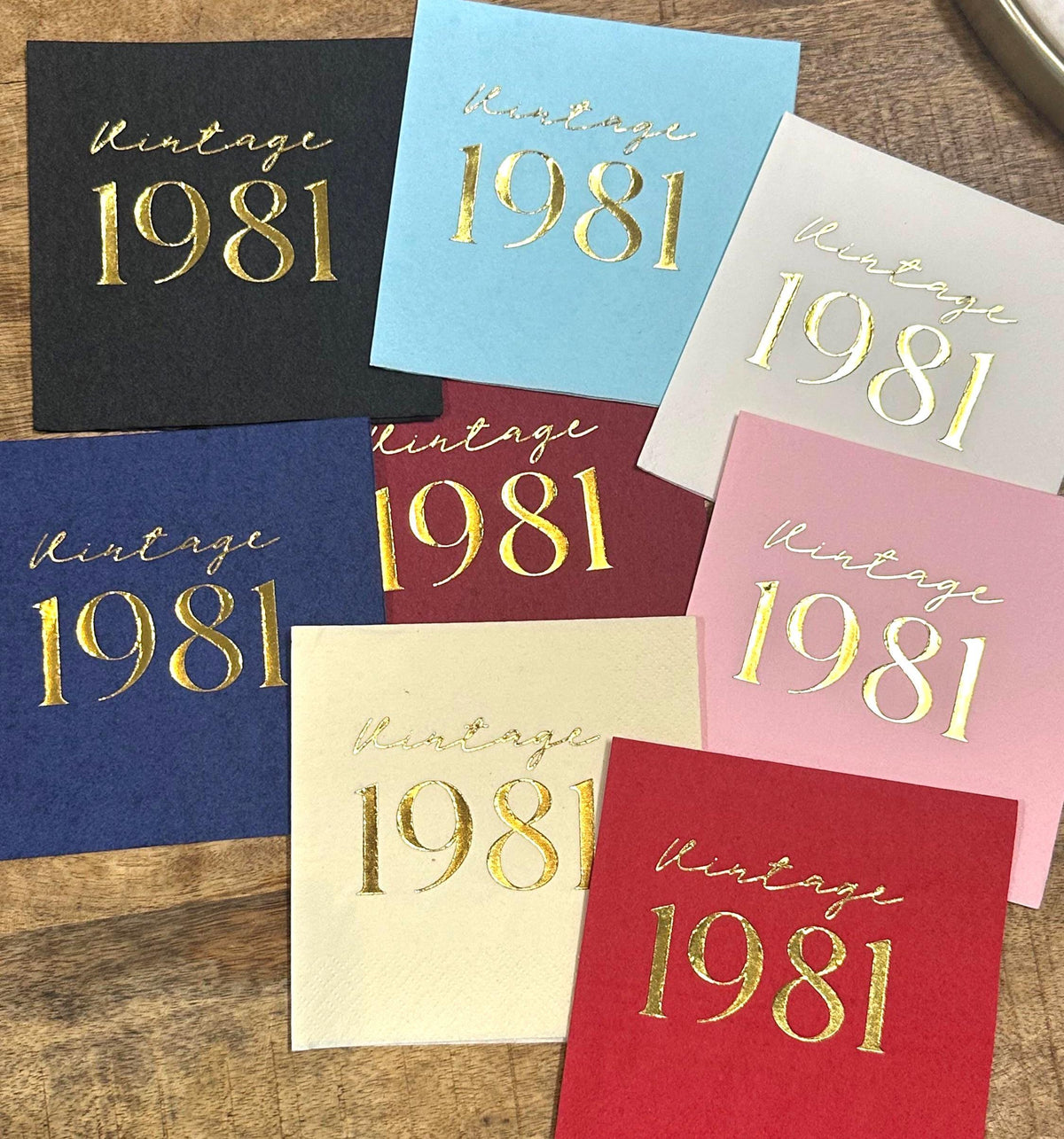 An arrangement of eight square paper napkins in various colors, each featuring the text Vintage 1981 in shiny gold foil.