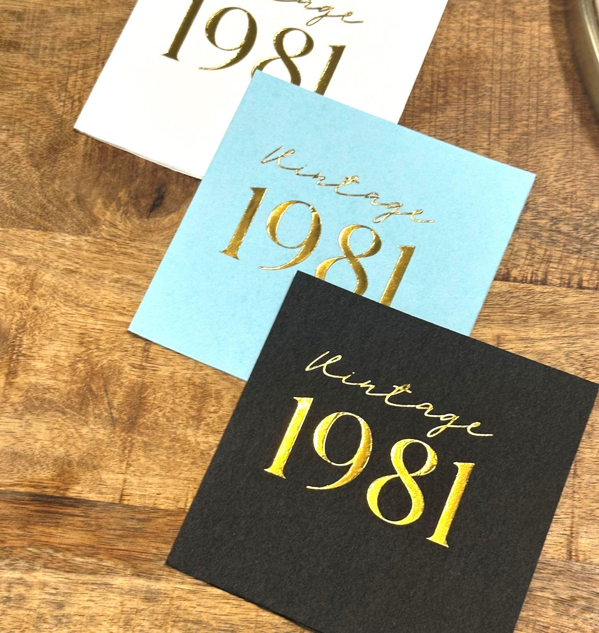 Three stacked square paper napkins in white, light blue, and black, each printed with Vintage 1981 in gold foil lettering.