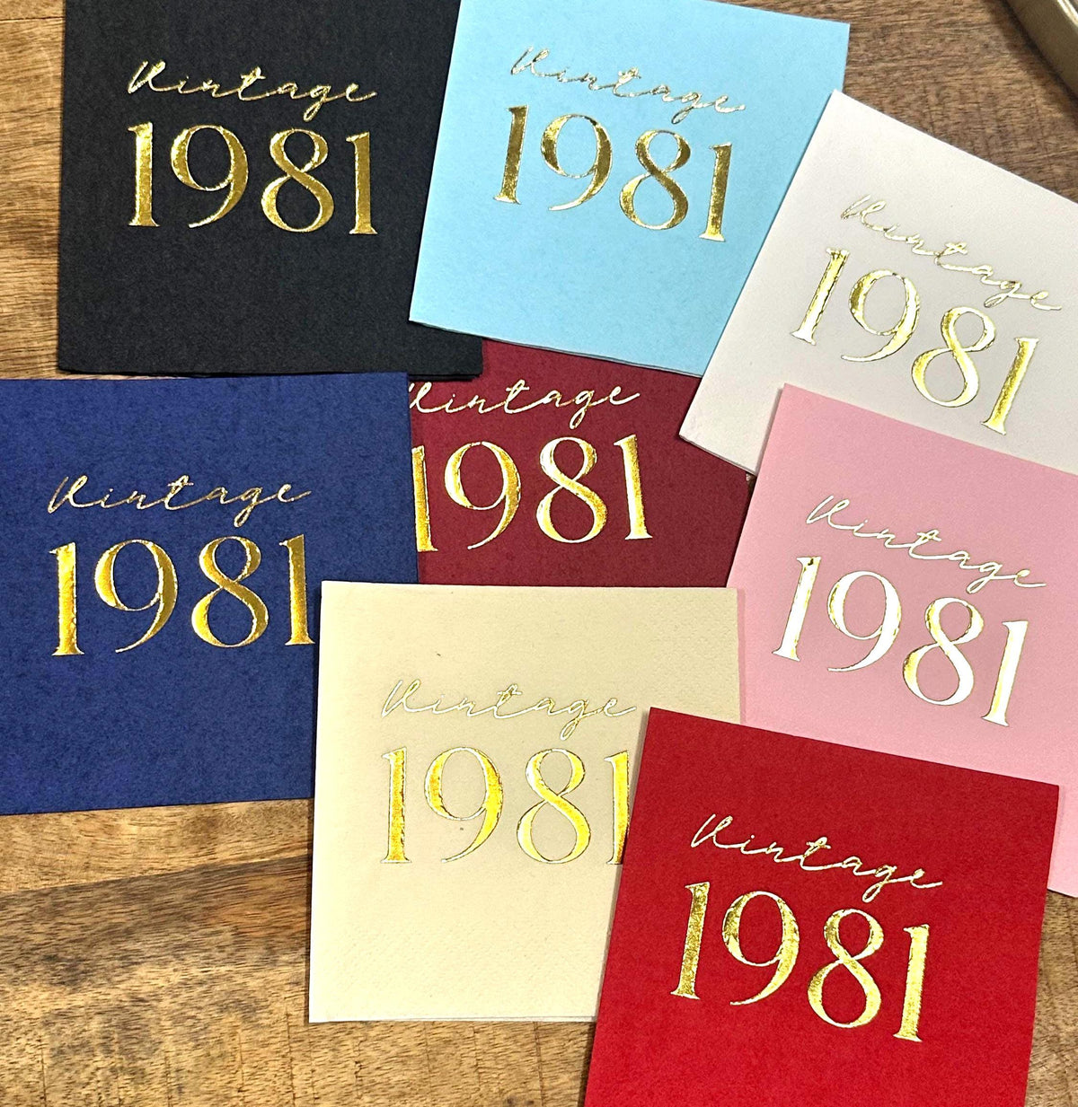 Seven square paper napkins in assorted colors, laid out in an overlapping pattern with gold foil Vintage 1981 text.