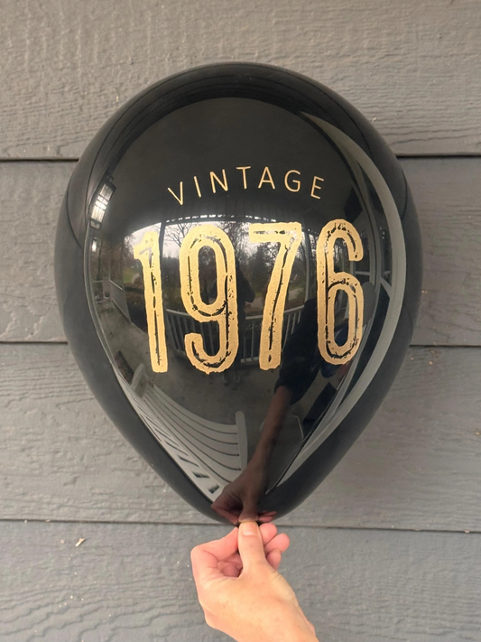 Black balloon with 'Vintage 1976' text held against a wooden surface