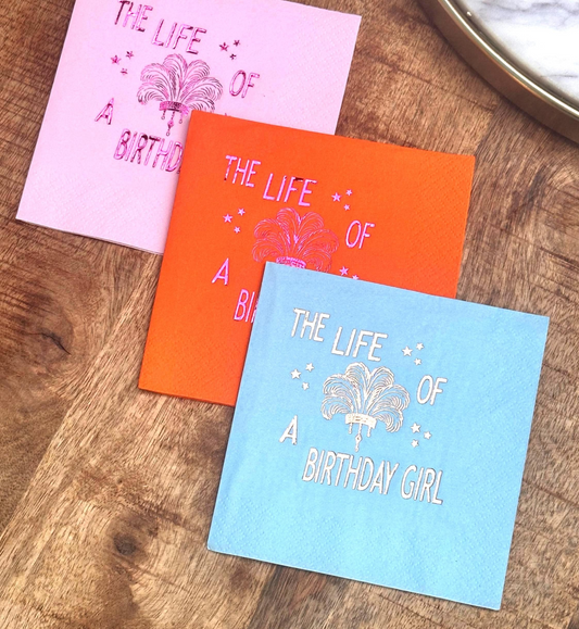 Foil-stamped “The Life of a Birthday Girl” cocktail napkins in pink, orange, blue, and white, featuring a feather plume and stars — perfect for Taylor Swift-inspired birthday parties or girly celebrations.